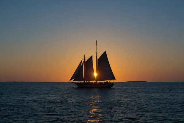 Two-masted schooner with passengers on deck, the setting sun between its sails off Key West