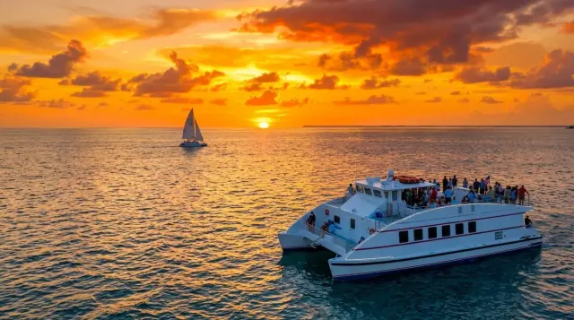 Catamaran carrying guests past a sailboat in an orange Key West sunset